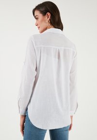 White long-sleeve shirt with textured fabric, buttoned cuffs, back pleat, and curved hem. Paired with blue jeans.