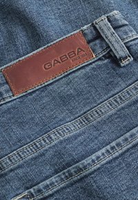 Džinsai, pasižymintys tamsiai mėlyna spalva, siuvinėjimu ir rudos odinės prekės ženklo etikete, kurioje rašoma "GABBA SINCE 1953".