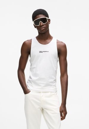 Fiatal férfi fehér ujjatlan felsőben, amelyen a "Karl Lagerfeld Jeans" felirat olvasható, fehér nadrágban és nagy fehér keretes napszemüvegben, kezei a zsebében.