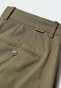 Poche arrière d'un pantalon vert olive avec fermeture par bouton et passants de ceinture, avec une petite étiquette de marque métallique rectangulaire.