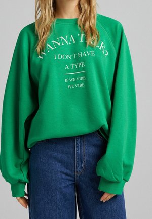 Personne portant un sweat-shirt vert avec le texte « TU VEUX PARLER ? JE N'AI PAS DE TYPE, SI ON VIBE, ON VIBE » et un jean bleu.
