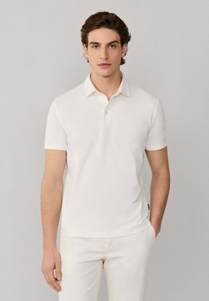 Joven con una camiseta polo blanca ajustada de manga corta y pantalones blancos, de pie contra un fondo liso de color gris claro.