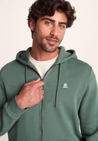Green zip-up hoodie featuring a logo patch, drawstring hood, and ribbed cuffs. Fabric appears soft with a smooth texture.