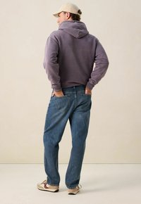 Man in een beige pet, paarse hoodie, blauwe jeans en beige sneakers met rode accenten, staande met zijn handen in de achterzakken.