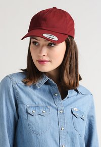 Cappellino da baseball bordeaux con una visiera tondeggiante e un design in tessuto. Presenta un'etichetta circolare con un logo. Indossato con una camicia in denim blu chiaro.