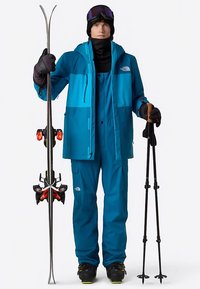 Turquoise skipak met verstelbare bandjes, gecombineerd met zwarte handschoenen en een nekwarmer. Houdt ski's en stokken vast, draagt een zwarte muts en skibril.