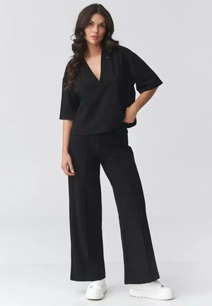 Kobieta o długich ciemnych włosach, ubrana w czarną bluzkę z dekoltem w serek, szerokie czarne spodnie oraz białe sneakersy na platformie, stojąca na jednolitym tle.