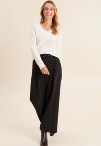 Weißer, strukturierter V-Ausschnitt-Pullover kombiniert mit hochtaillierten, schwarzen weit geschnittenen Hosen mit einer runden Gürtelschnalle. Schwarze Stiefeletten runden den Look ab.