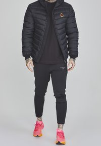 Černá péřová bunda s kapucí a vyšitým znakem, kombinovaná s přiléhavými černými kalhotami a živě růžovými sportovními botami.