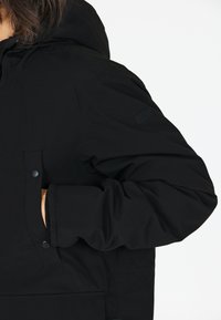 Sort isoleret jakke med hætte, trykknaplommer og et tekstureret stof med et logo-patch på ærmet.