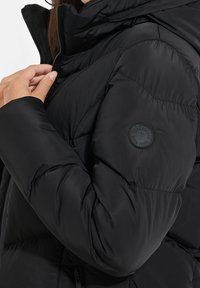 Veste matelassée noire avec col montant, manches retroussées et écusson rond en caoutchouc sur l'épaule ; texture lisse et brillante.