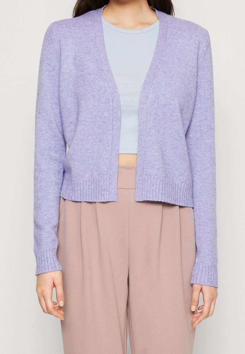 Femme portant un cardigan lavande ouvert sur un haut court bleu clair et un pantalon beige taille haute, debout devant un fond uni.