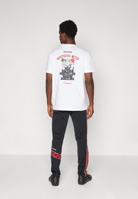 White cotton t-shirt featuring a graphic print of Manchester United and text on the back. The model wears black pants with red accents.