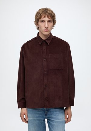 Young man with curly blond hair wearing a dark brown corduroy button-up shirt and light blue jeans against a plain background.