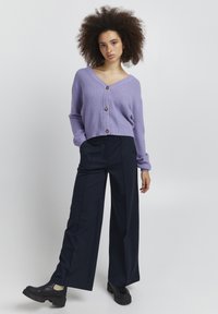 Kvinne med krøllete hår iført en lavendel knappet cardigan, vide marineblå bukser og svarte kraftige sko som står mot en ensfarget bakgrunn.