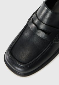 Black leather loafer with a square toe, featuring stitched detailing and a strap across the instep. Smooth texture with minimal sheen.