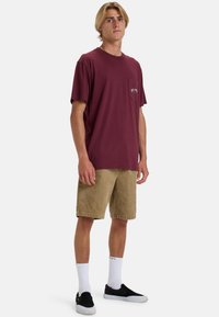 Maroons T-shirt i bomuld med en brystlomme, parret med tanfarvede shorts. Stylet med hvide sokker og sorte slip-on sko. En simpel, afslappet stil.