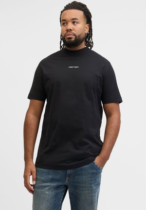Mand med dreadlocks iført sort t-shirt med korte ærmer med teksten "CORE™ DEPT." og blå jeans, står med den ene hånd i lommen mod en ensfarvet baggrund.