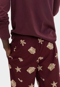 Pantalones de pijama color burdeos con patrones de temática navideña en beige que incluyen hombres de jengibre, árboles y bastones de caramelo. Tejido suave y elástico.