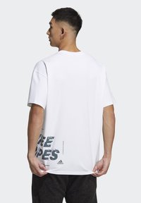 T-shirt bianco in cotone con vestibilità oversize, maniche corte e stampa grafica grigia sulla parte bassa della schiena. Logo Adidas ben visibile.