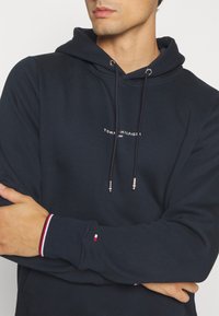 Felpa navy in morbido tessuto con logo frontale, cappuccio con coulisse e dettagli in argento, e polsini a righe con dettagli in rosso, bianco e blu.