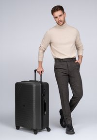 Valigia rigida nera con design a onde testurizzate, manico estensibile, ruote lisce e maniglia laterale; modello in maglione beige e pantaloni grigi.