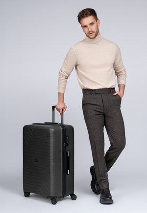 Schwarzer Hartschalenkoffer mit strukturiertem Wellenmuster, ausziehbarem Griff, glatten Rollen und seitlichem Griff; Modell in beigem Pullover und grauen Hosen.