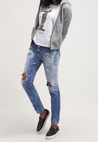 Ljusgrå zip-up hoodie, vit t-shirt med svart text, slitna blå jeans med knäskador och svarta skor utan snörning.