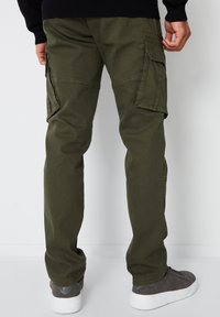Olivově zelené cargo kalhoty se dvěma bočními kapsami, se střihovými nohavicemi a odolnou bavlněnou tkaninou. Nosené s šedými teniskami.