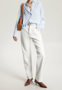 Camicia azzurro chiaro con bottoni, pantaloni bianchi a gamba dritta, scarpe con tacco marroni e borsa a tracolla marrone. Gli accessori includono collane a più strati.