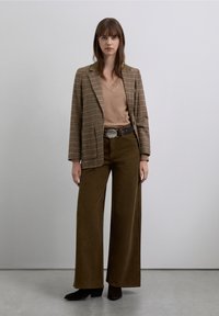Blazer de cuadros marrón, suéter beige con cuello en V, pantalones de pana anchos de pierna en oliva y botines negros. Hebilla de cinturón plateada que acentúa el conjunto.