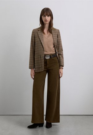 Blazer de cuadros marrón, suéter beige con cuello en V, pantalones de pana anchos de pierna en oliva y botines negros. Hebilla de cinturón plateada que acentúa el conjunto.