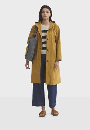 Kvinde iført en sennepsgul hættetrøje, stribet sweater, vide jeans, brune sandaler og bærer en stribet skuldertaske.