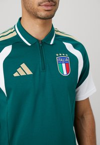 Mann med grønn Italia fotballtrøye med glidelås krage, hvite paneler på ermene, Adidas-logo og Italias landslagsmerke med fire stjerner.