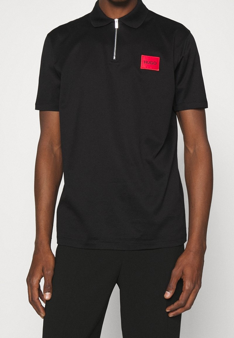 Polo noir en coton avec un col zippé, présentant un logo rectangulaire rouge sur la poitrine gauche. Design simple à manches courtes.