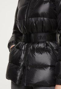 Sort puffert jakke med blank finish, vandret quiltet tekstur, to lynlåslommer og et kontrastfarvet sort logo-bælte i taljen.