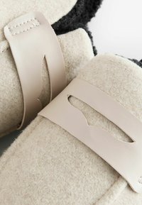 Filzslipper mit Lederriemen in Creme und Schwarz, mit einem ausgeschnittenen Design an den Riemen und einem strukturierten, weichen Material.