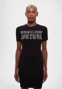 Čierne priliehavé šaty s krátkymi rukávmi, s logom "Versace Jeans Couture" v metalicky striebornom písme cez prsia. Hladká textúra.