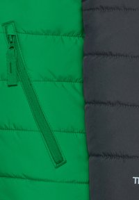 Gepolsterte Jacke mit grünen und schwarzen Einsätzen, horizontalen Nähten und einer grünen Stofftasche mit Reißverschluss. Einfache Markenkennzeichnung.