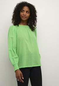 Blouse verte texturée avec des manches bouffantes, un col rond et des poignets smockés. Portée avec un pantalon en denim foncé.