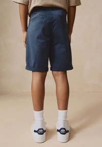 Pantalones cortos de algodón azul con dobladillos enrollados, dos bolsillos traseros, combinados con zapatillas blancas y calcetines blancos acanalados, mostrando un fondo neutro.