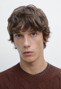 Persoon met middellang, golvend bruin haar. Draagt een bruin kabelgebreide trui over een donkerder bruine shirt. Minimalistische achtergrond.