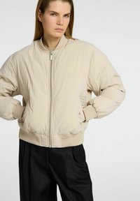 Una modella con capelli lunghi indossa una giacca bomber beige chiaro, posando con sicurezza su uno sfondo neutro, mettendo in risalto il design della giacca.