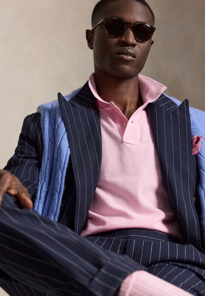 Uomo che indossa un completo blu scuro a righe, una polo rosa, un maglione azzurro chiaro drappeggiato sulle spalle e occhiali da sole scuri seduto con le gambe incrociate.
