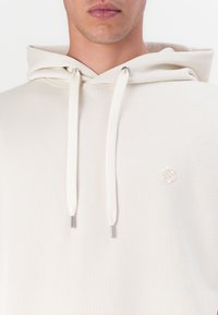 Crème kleurige hoodie van een textuurstof, met een klein geborduurd logo op de borst en zilverkleurige stoppers aan de koorden.