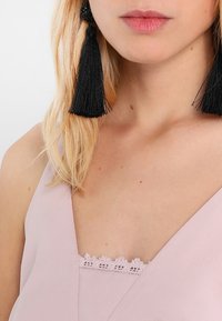 Light pink sleeveless top featuring a lace trim neckline, paired with large black tassel earrings. Smooth fabric with a subtle sheen.