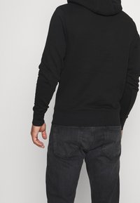 Hombre con una sudadera negra y jeans desgastados negros visto desde atrás contra un fondo claro y simple.