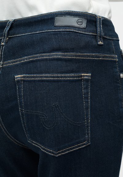 Σκούρο μπλε τζιν με υφή επιφάνειας, που διαθέτει πίσω τσέπη με ραμμένες λεπτομέρειες και ορθογώνιο μαύρο λογότυπο στη μέση.