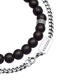 Bracciale con perle nere opache, catena d'argento, un elemento decorativo in argento e una targhetta incisa con "Kuzoi." Texture liscia in tutto.