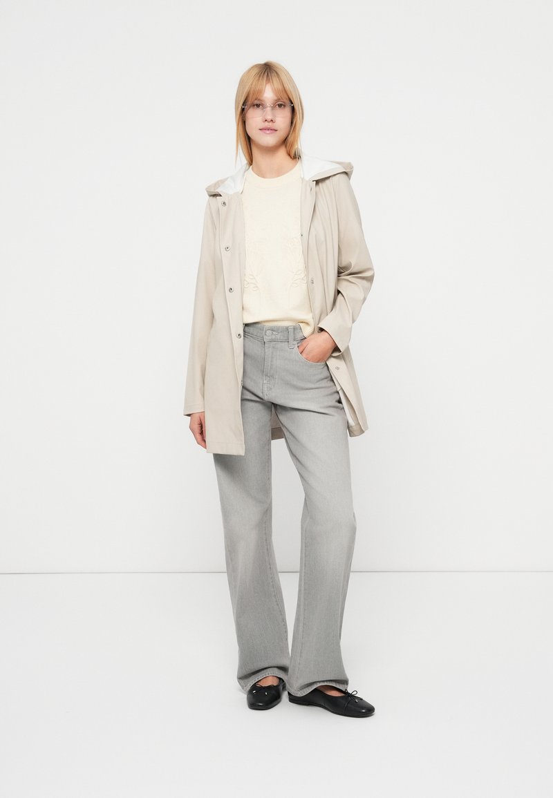 Chaqueta impermeable beige con capucha blanca, suéter ligero y pantalones acampanados gris claro, combinados con zapatos planos negros, posando frente a un fondo blanco.